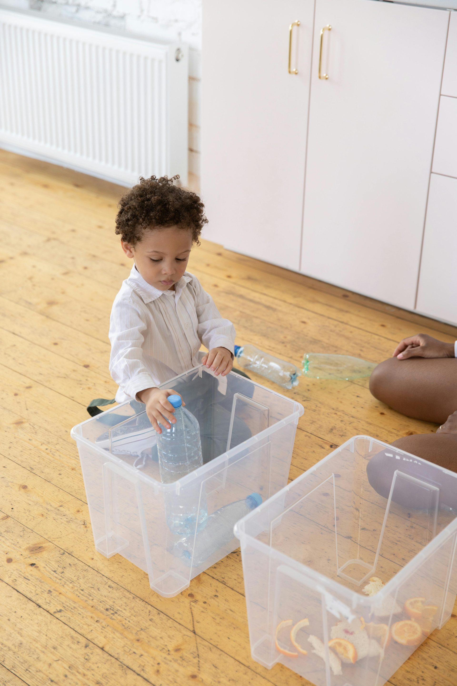 Young child sorting plastic bottles into containers in a kitchen setting.