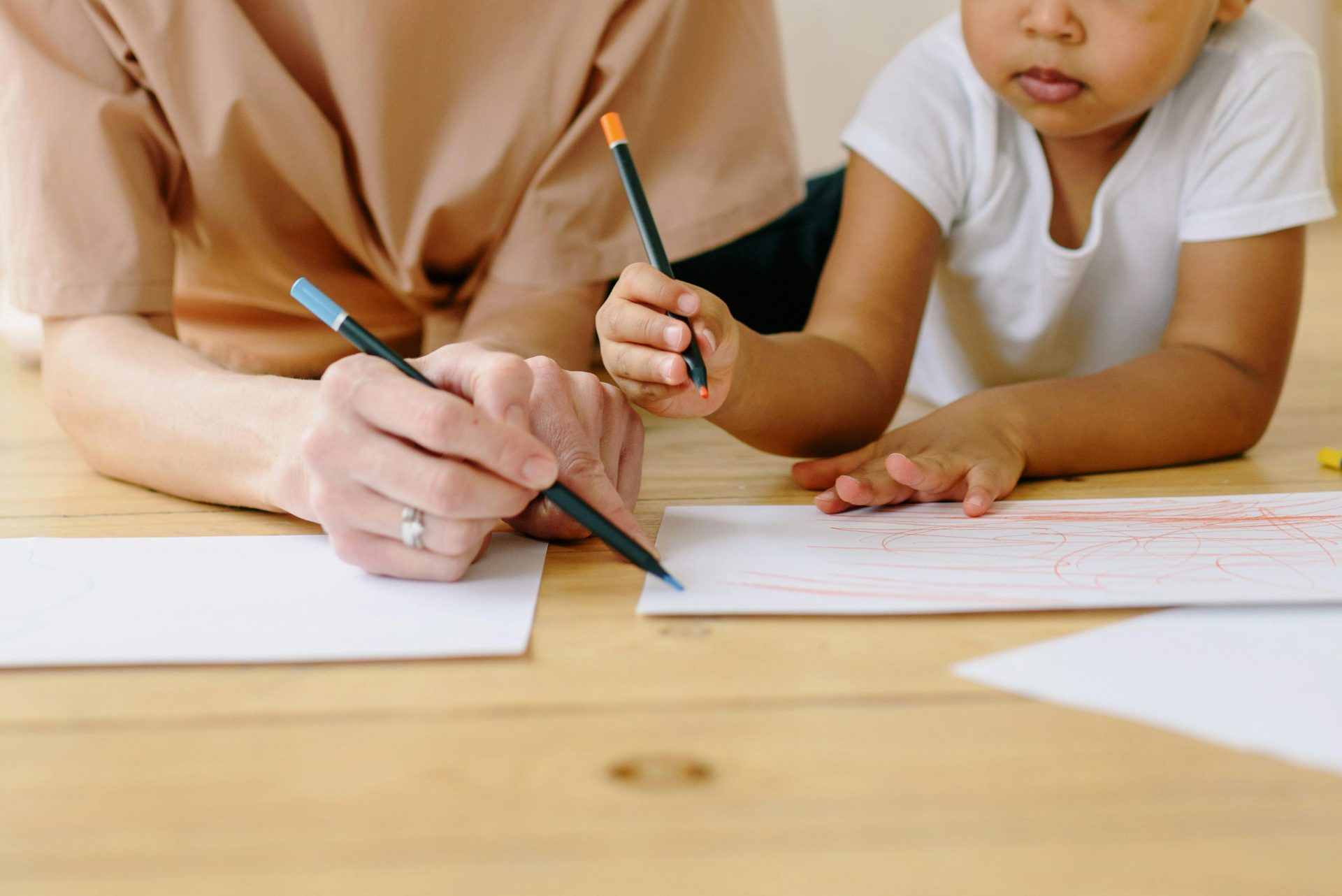 A woman and child drawing with color pencils on white paper around a table indoors.