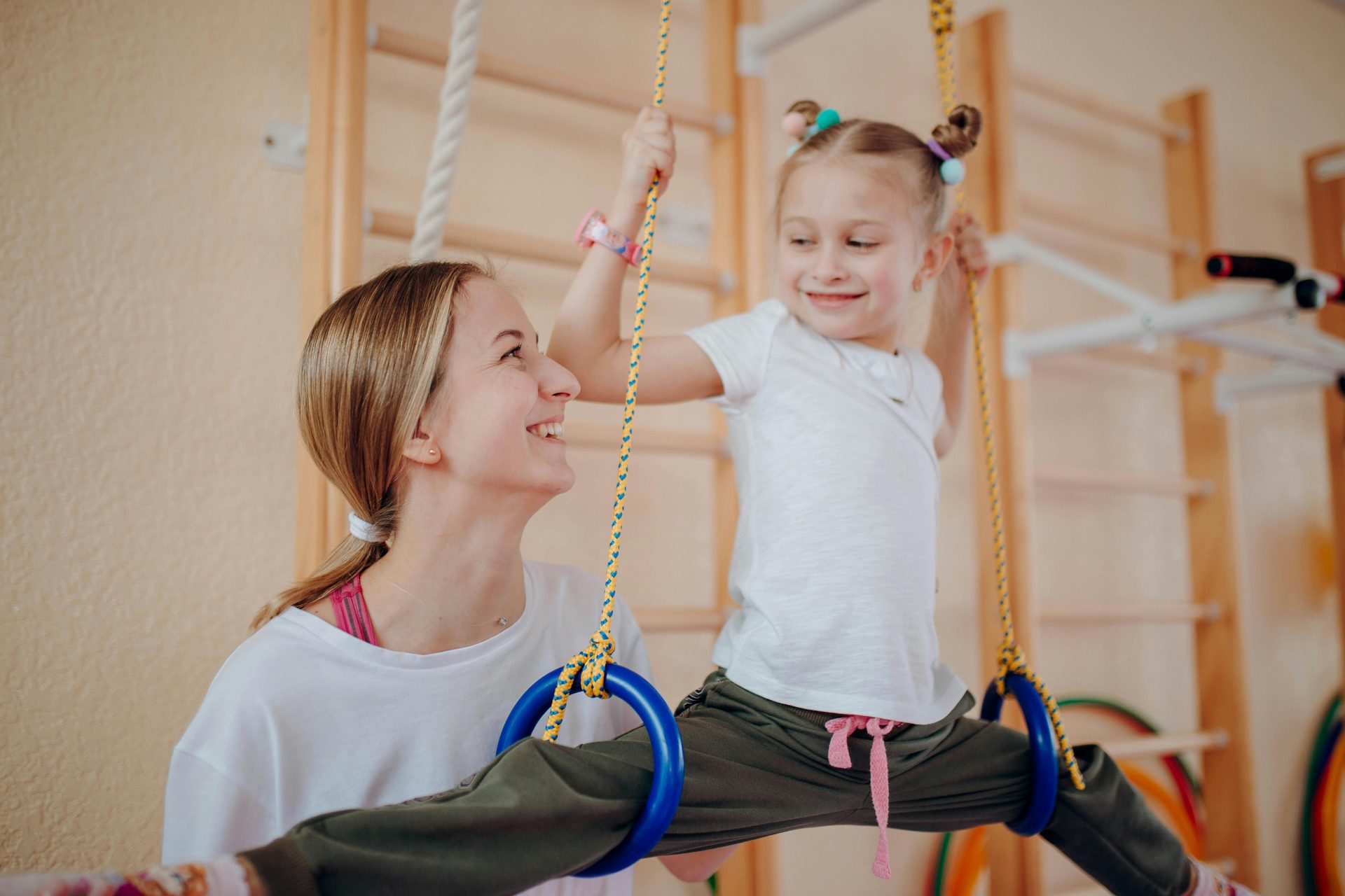 Smiling mother teaches her daughter gymnastics on rings indoors, fostering bonding and flexibility.