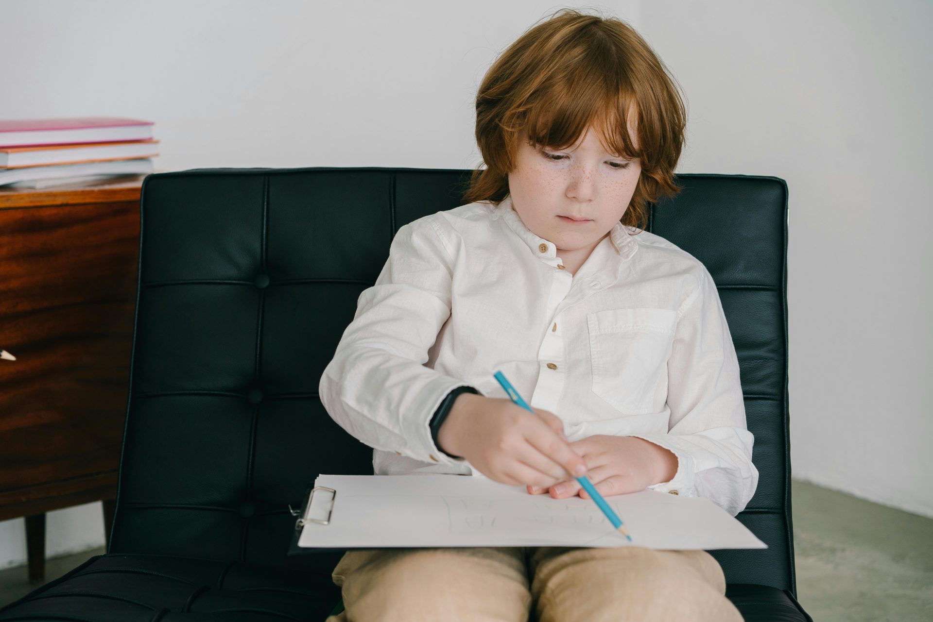Child with red hair concentrates on drawing with colored pencils while sitting on a black chair.